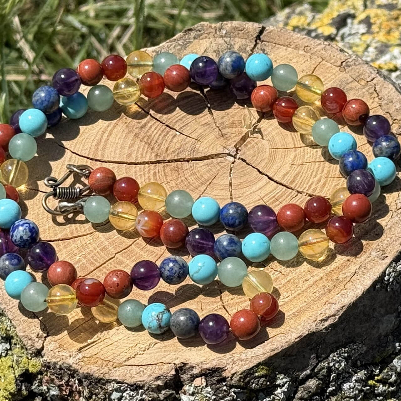 7 Chakra Necklace displayed on a wooden block in a garden with green grass background